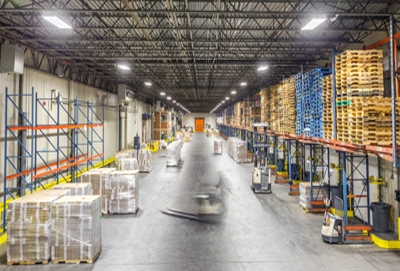 Warehouse with rows of pallet racks and forklifts. The floor appears to be concrete with some wear and tear.